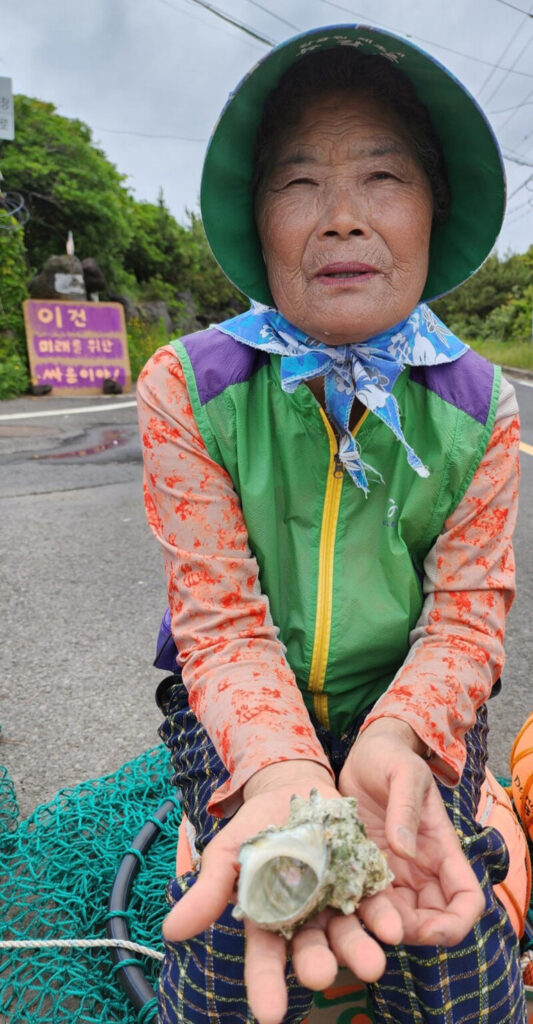 A haenyeo wearing a green half sleeve jacket, green hat holds a hollow conch shell to demonstrate the state of pollution in the sea in Woljeong-ri.
© Provided by The Hankyoreh